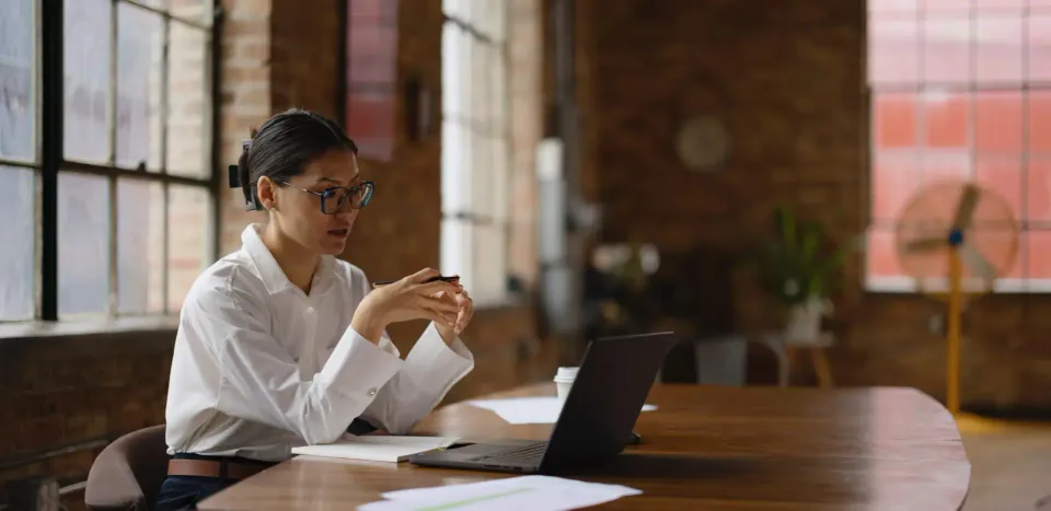 Une professionnelle installée à une grande table relit un document devant son ordinateur portable